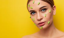 Artistic portrait of a woman with green capsules on her face against a yellow background.